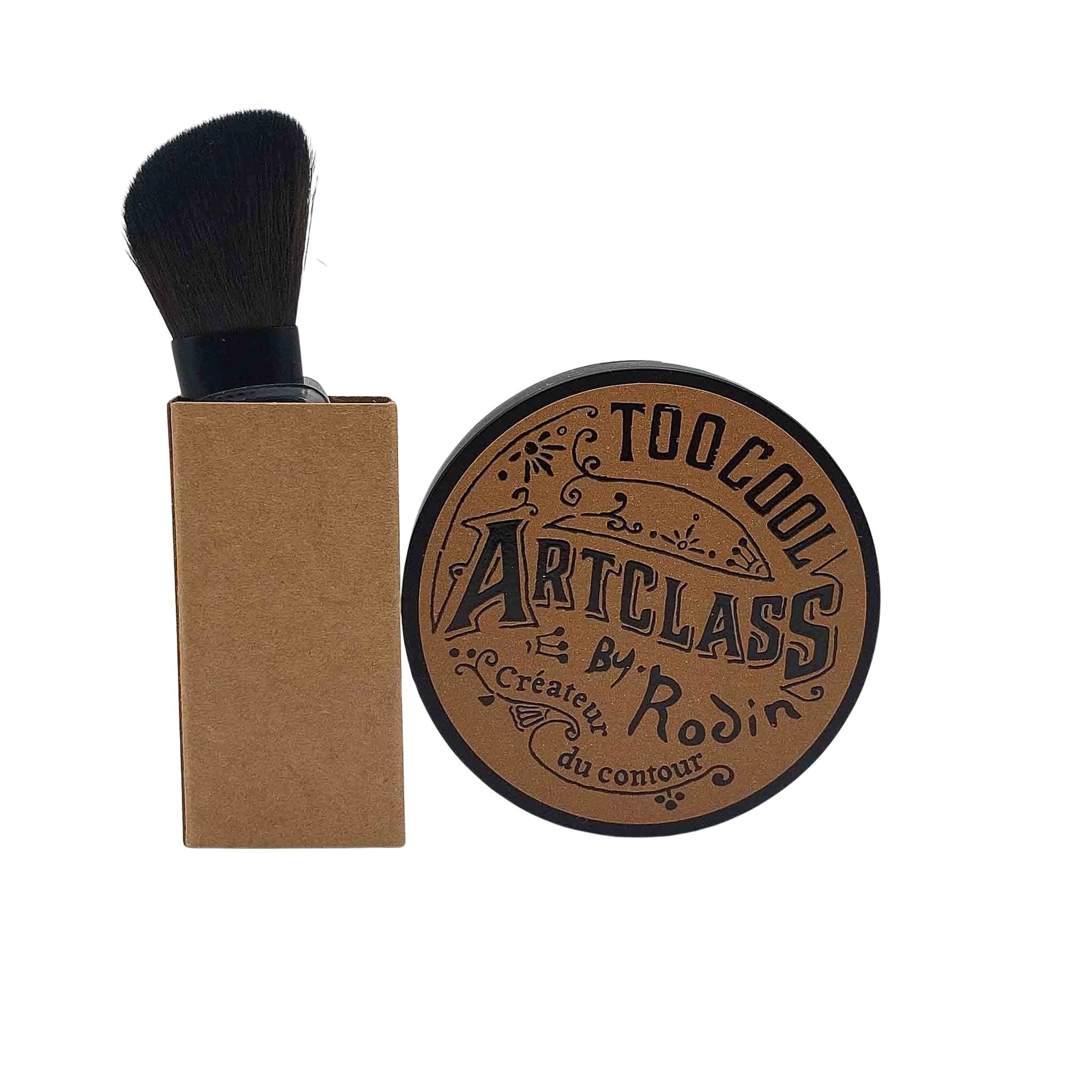 Bronze round Artclass by Rodin shading compact with ornate typography, beside a rectangular kraft brush with black bristles.